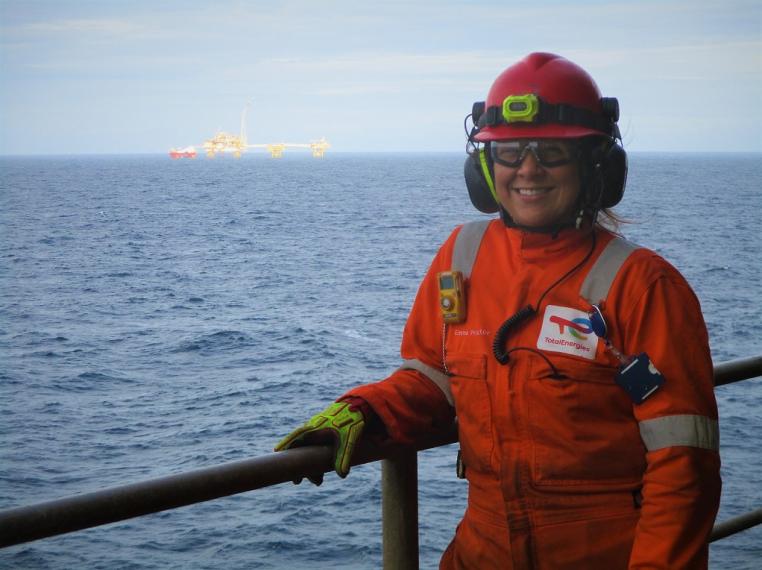 Woman standing in orange overalls with sea and oil platform in background
