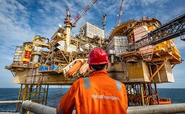 Man wearing red helmet and orange overalls looking up to an oil rig