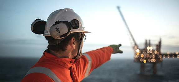 Man wearing white helmet and orange overalls pointing to an oil rig in the distance